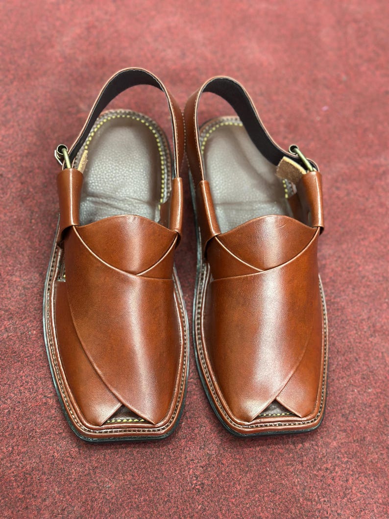 K&ouml;nnte beinhalten: Ein Paar braune Ledersandalen. Die Sandalen haben ein unverwechselbares Design mit einem Riemen um den Kn&ouml;chel und einer quadratischen Zehenpartie. Das Leder hat eine gl&auml;nzende Oberfl&auml;che. Die Sohlen sind schwarz.