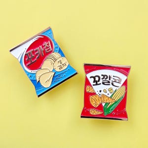 May include: Two enamel pins in the shape of snack food packets. One is blue and red with the Korean text "포카칩" and potato chip images. The other is red and white with the Korean text "꼬깔콘" and corn images.