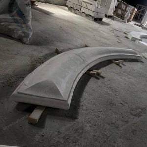 Könnte beinhalten: Ein großes, gebogenes architektonisches Steinelement, wahrscheinlich eine Leiste oder ein Gesims, in hellgrauer Farbe. Das Stück wird von kleinen Holzblöcken auf einem Betonboden getragen. Weitere Steinelemente sind im Hintergrund zu sehen.