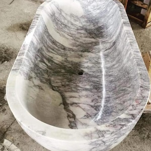 May include: An oval-shaped freestanding bathtub carved from natural stone with a polished finish. The basin features a distinct white base color with prominent, swirling grey and purple veining throughout the interior and exterior surfaces.