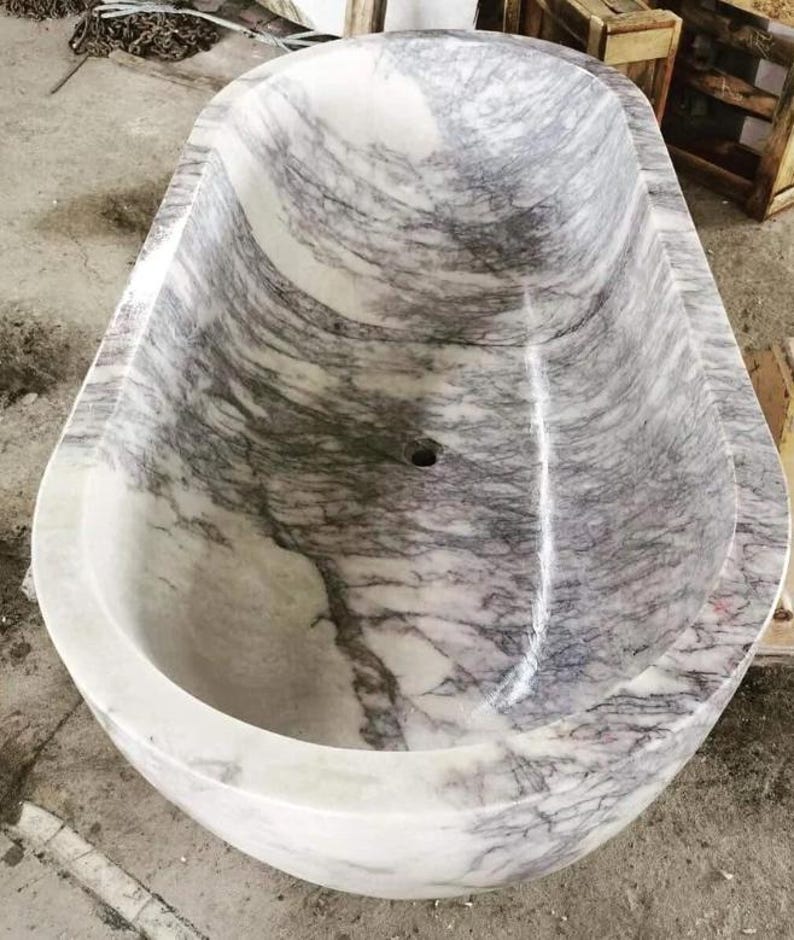 May include: An oval-shaped freestanding bathtub carved from natural stone with a polished finish. The basin features a distinct white base color with prominent, swirling grey and purple veining throughout the interior and exterior surfaces.
