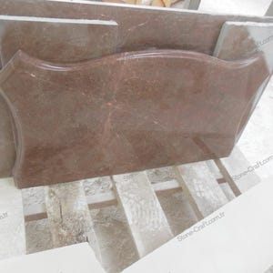 May include: A collection of reddish-brown marble slabs, including a large, uniquely shaped piece with curved edges. The marble has natural veining and is stacked on a wooden pallet. The pieces vary in size and shape, suggesting a selection for construction or design.