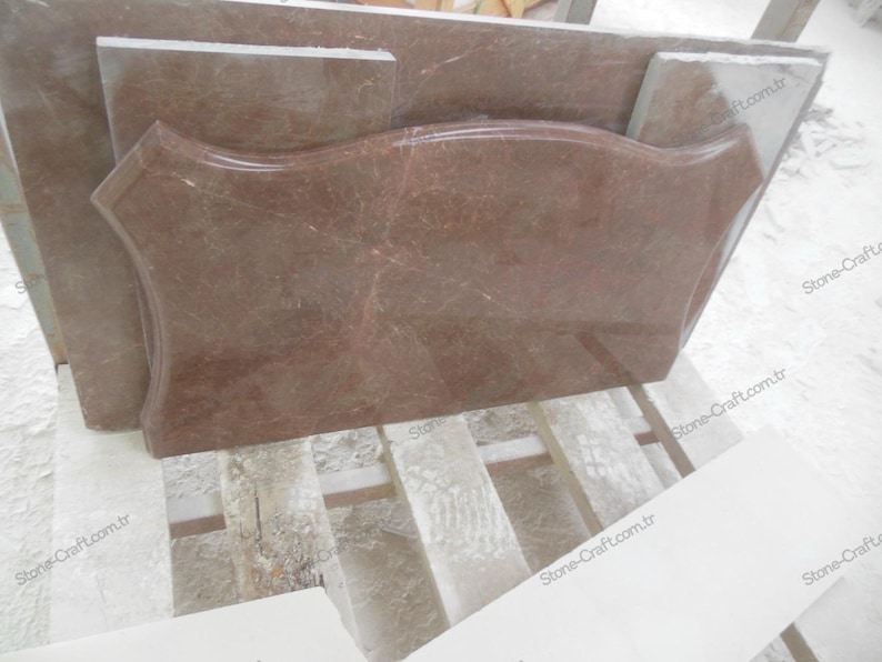 May include: A collection of reddish-brown marble slabs, including a large, uniquely shaped piece with curved edges. The marble has natural veining and is stacked on a wooden pallet. The pieces vary in size and shape, suggesting a selection for construction or design.