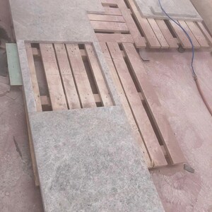 May include: Gray stone slabs of varying sizes are arranged on wooden pallets. The slabs have a textured surface and are likely for construction or design purposes. The scene is indoors, with additional rolled materials visible in the background.
