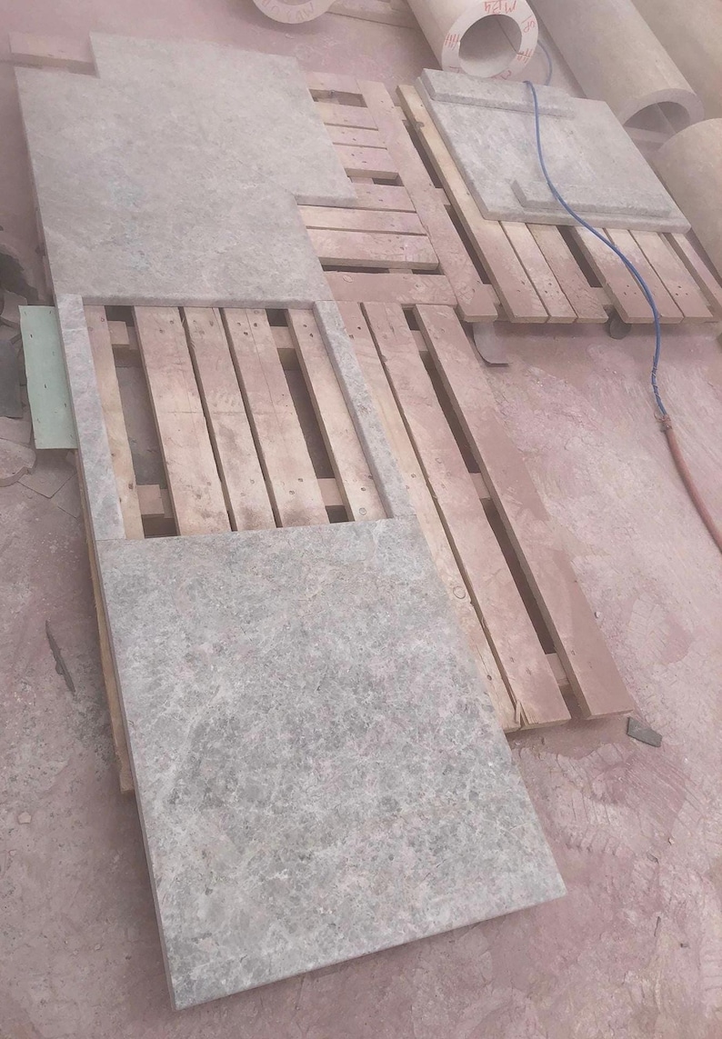 May include: Gray stone slabs of varying sizes are arranged on wooden pallets. The slabs have a textured surface and are likely for construction or design purposes. The scene is indoors, with additional rolled materials visible in the background.