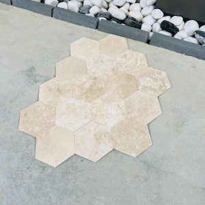 May include: A collection of beige hexagonal tiles arranged on a concrete surface. The tiles have a natural stone appearance with subtle variations in color and texture. The tiles are surrounded by a border of gray concrete and white pebbles.