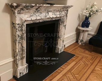 Könnte beinhalten: Ein Kaminumbau aus weißem Marmor mit ausgeprägter dunkelgrauer Maserung, bestehend aus einem rechteckigen Sims und einem schwarzen Kamineinsatz. Der Text STONE-CRAFT.com.tr ist in der Mitte der Kaminfront zu sehen.