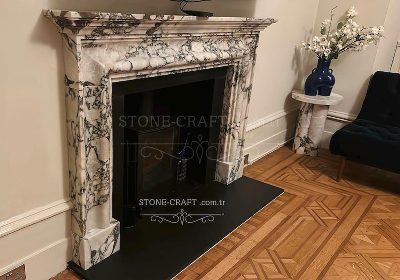 Puede incluir: Una repisa de chimenea hecha de m&aacute;rmol blanco con vetas gris oscuro prominentes, que presenta un estante rectangular y un hogar interior negro. El texto STONE-CRAFT.com.tr es visible en el centro de la estructura de la chimenea.