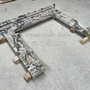 Puede incluir: Una repisa de chimenea hecha de m&aacute;rmol blanco con vetas pronunciadas en color morado oscuro y negro. La estructura presenta un estante superior horizontal y dos pilares laterales verticales con bordes moldeados. El texto STONE-CRAFT.com.tr es visible.