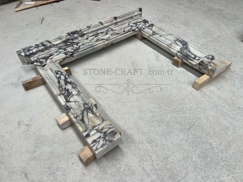 Puede incluir: Una repisa de chimenea hecha de m&aacute;rmol blanco con vetas pronunciadas en color morado oscuro y negro. La estructura presenta un estante superior horizontal y dos pilares laterales verticales con bordes moldeados. El texto STONE-CRAFT.com.tr es visible.