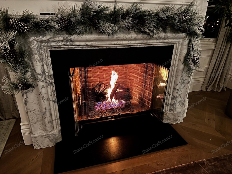 以下が含まれることがあります： 白い大理石の暖炉の周囲には、黒い金属製のインサートがあり、内部で火が燃えています。暖炉の内部は赤いレンガでできており、上部には松ぼっくりが付いた長い霜降りの緑色の松のガーランドが飾られています。