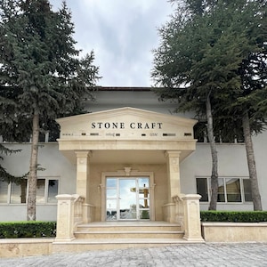 May include: Exterior view of a building with the text "STONE CRAFT" above the entrance. The building has a light beige facade, a glass door, and is flanked by tall evergreen trees. The sky is overcast.