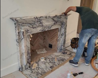 Puede incluir: Una chimenea de mármol blanco con vetas pronunciadas en color púrpura oscuro y gris. La estructura presenta un diseño clásico de repisa con columnas verticales que flanquean la abertura, revestida con azulejos en espiga de color marrón claro.