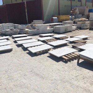 May include: Outdoor shot of various stone slabs and blocks, likely for construction or landscaping. The slabs are light gray and rest on wooden pallets. Other stone elements, including rectangular blocks and larger pieces, are visible in the background.