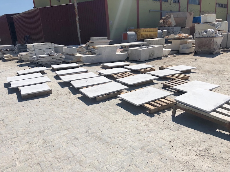 May include: Outdoor shot of various stone slabs and blocks, likely for construction or landscaping. The slabs are light gray and rest on wooden pallets. Other stone elements, including rectangular blocks and larger pieces, are visible in the background.