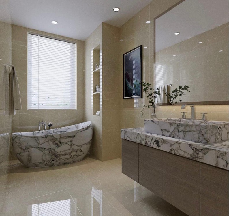 May include: A bathroom interior featuring a freestanding bathtub and a rectangular sink basin, both crafted from white marble with prominent grey veining. The vanity has a wooden cabinet base, and the walls are covered in large beige tiles.