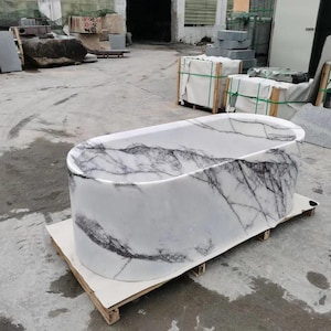 May include: A freestanding oval bathtub carved from white stone with prominent dark gray and black veining throughout the exterior and interior surfaces. The tub has a smooth, polished finish and is positioned on a wooden pallet for transport.