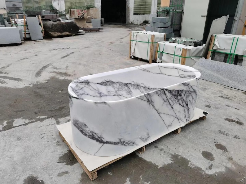 May include: A freestanding oval bathtub carved from white stone with prominent dark gray and black veining throughout the exterior and interior surfaces. The tub has a smooth, polished finish and is positioned on a wooden pallet for transport.