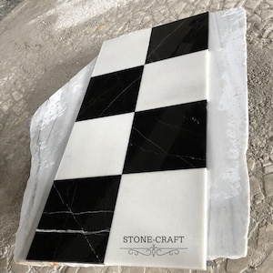 Op de afbeelding: Een rechthoekige marmeren tegel met een dambordpatroon van zwarte en witte vierkanten. De tegel heeft een gepolijste afwerking en de tekst "STONE-CRAFT" is zichtbaar. De tegel ligt op een grotere, ruwe marmeren plaat.