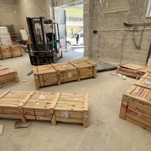 Puede incluir: Varias cajas de env&iacute;o rectangulares de madera con listones horizontales y bandas met&aacute;licas, etiquetadas con el texto Stone Craft, dispuestas sobre un suelo de hormig&oacute;n en un almac&eacute;n con una carretilla elevadora al fondo cerca de una puerta.