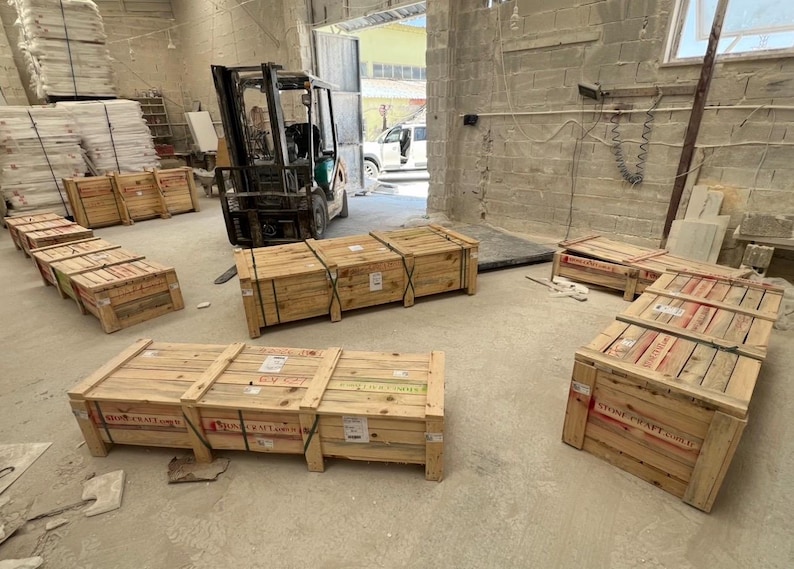 Puede incluir: Varias cajas de env&iacute;o rectangulares de madera con listones horizontales y bandas met&aacute;licas, etiquetadas con el texto Stone Craft, dispuestas sobre un suelo de hormig&oacute;n en un almac&eacute;n con una carretilla elevadora al fondo cerca de una puerta.