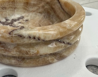 May include: A round bowl crafted from polished honey-colored onyx stone, featuring natural translucent layers, swirling cream and tan patterns, and distinct dark brown mineral veins throughout the smooth, thick, and rounded rim and interior surface.