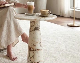 Könnte beinhalten: Ein runder Beistelltisch aus weißem Marmor mit ausgeprägter brauner Maserung und einem konischen Sockel. Auf der Tischplatte stehen ein Glas Eiskaffee mit Strohhalm sowie eine Keramiktasse mit heißem Kaffee, jeweils auf passenden Untertassen.