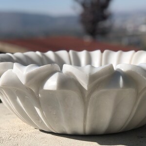 May include: A white marble bowl with a decorative, carved design resembling a flower. The bowl is smooth and polished, with a delicate, intricate pattern.