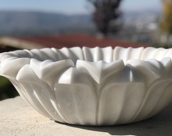 May include: A white marble bowl with a decorative, carved design resembling a flower. The bowl is smooth and polished, with a delicate, intricate pattern.