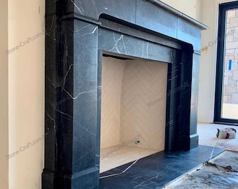 Könnte beinhalten: Ein großer Kamin aus schwarzem Marmor mit weißen Adern. Der Kamin hat eine rechteckige Öffnung und einen breiten Mantel. Das Innere des Kamins ist mit weißen Fischgrätfliesen ausgekleidet. Der Raum hat helle Wände.
