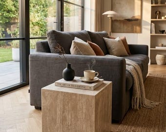 Könnte beinhalten: Ein beigefarbener, würfelförmiger Beistelltisch aus Naturstein mit poröser Textur. Darauf liegen ein Buch mit dem Titel The Art of Travertine, eine kleine schwarze Vase mit einem getrockneten Zweig sowie eine dampfende Tasse auf einer Untertasse.