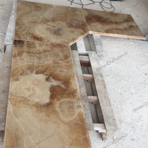 Könnte beinhalten: Eine große, rechteckige Platte aus poliertem Onyxstein mit einer warmen, beige-braunen Farbpalette. Der Stein weist natürliche Maserungen und Muster auf und ist in mehrere Teile geschnitten. Die Teile sind auf einer Holzkonstruktion angeordnet.