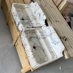 May include: Two rectangular marble farmhouse sinks featuring a white base with prominent grey and brown veining. Each sink has a fluted exterior front and a high back splash. A yellow measuring tape is placed on the wooden crate nearby.