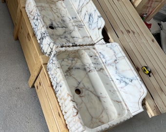 May include: Two rectangular marble farmhouse sinks featuring a white base with prominent grey and brown veining. Each sink has a fluted exterior front and a high back splash. A yellow measuring tape is placed on the wooden crate nearby.