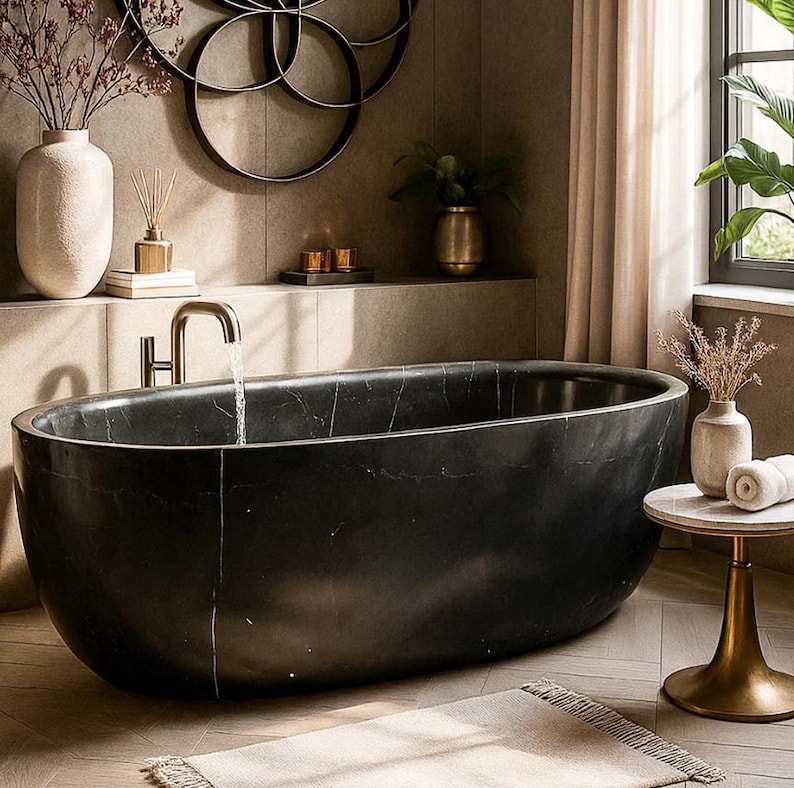 May include: A freestanding oval bathtub made of black stone with subtle white veining. A brushed metal faucet is mounted on the edge, with water flowing into the basin. The tub is positioned in a room with neutral-toned walls and decor.