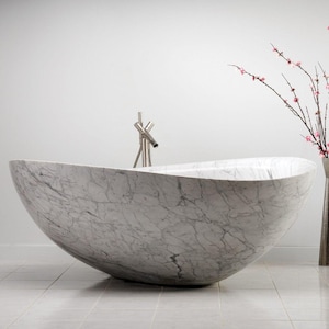 May include: A freestanding oval bathtub crafted from white marble with prominent grey veining. The tub features a smooth, curved silhouette and is paired with a tall, slender metallic faucet fixture positioned at the center of the rim.