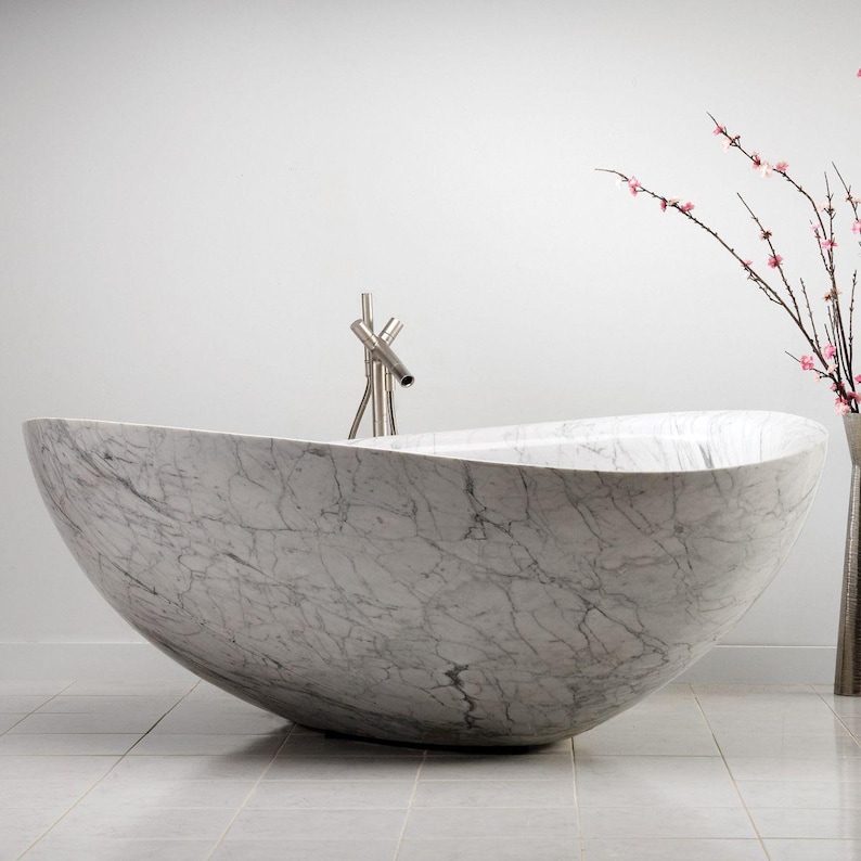 May include: A freestanding oval bathtub crafted from white marble with prominent grey veining. The tub features a smooth, curved silhouette and is paired with a tall, slender metallic faucet fixture positioned at the center of the rim.