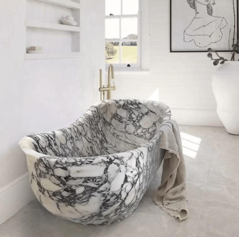 May include: A freestanding oval bathtub crafted from white marble with prominent black veining. The tub features a smooth, polished finish and is draped with a beige textured towel. A gold-toned faucet is mounted on the wall behind the basin.