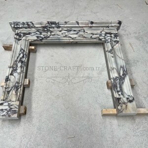 Puede incluir: Una repisa de chimenea hecha de m&aacute;rmol blanco con vetas prominentes de color morado oscuro y negro. La estructura presenta un estante superior rectangular y dos pilares laterales verticales con molduras decorativas. El texto STONE-CRAFT.com.tr es visible.