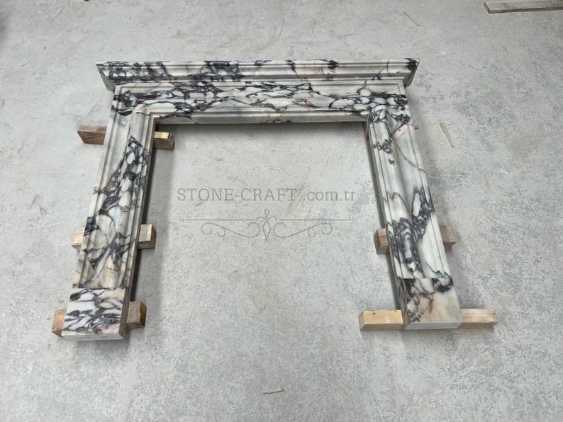 Puede incluir: Una repisa de chimenea hecha de m&aacute;rmol blanco con vetas prominentes de color morado oscuro y negro. La estructura presenta un estante superior rectangular y dos pilares laterales verticales con molduras decorativas. El texto STONE-CRAFT.com.tr es visible.