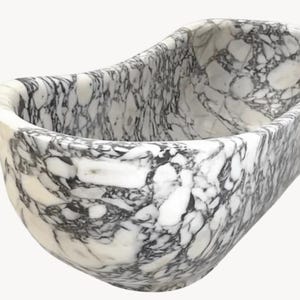 May include: A freestanding bathtub carved from white marble with prominent, irregular black veining throughout the entire surface. The tub features a smooth, rounded, elongated oval shape with high, curved walls and a deep, open interior basin.