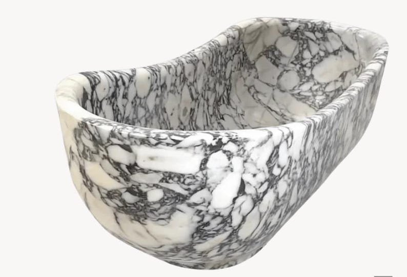 May include: A freestanding bathtub carved from white marble with prominent, irregular black veining throughout the entire surface. The tub features a smooth, rounded, elongated oval shape with high, curved walls and a deep, open interior basin.