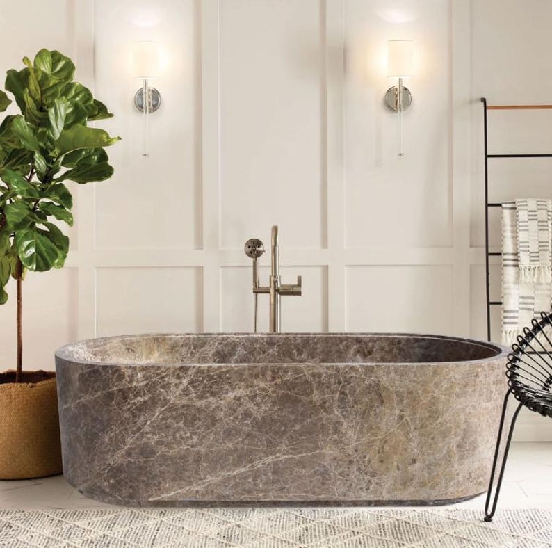 May include: A freestanding oval bathtub crafted from brown marble with natural veining patterns. The tub features a smooth, polished finish and is paired with a tall, brushed nickel floor-mounted faucet with a handheld shower attachment.