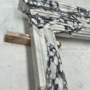 Puede incluir: Una repisa de chimenea de m&aacute;rmol blanco con vetas de color p&uacute;rpura oscuro, acabado pulido y bordes moldeados. La estructura de piedra muestra un patr&oacute;n de alto contraste con vetas violetas intensas sobre un fondo blanco. Texto: STONE-CRAFT.com.tr.
