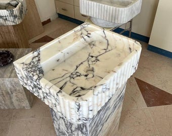 Könnte beinhalten: Ein rechteckiges Standwaschbecken aus weißem Marmor mit markanter dunkelgrauer und schwarzer Äderung. Das Becken verfügt über ein geriffeltes Außendesign an der Vorderseite und den Seiten sowie ein zentrales Abflussloch und eine glatte Oberfläche.