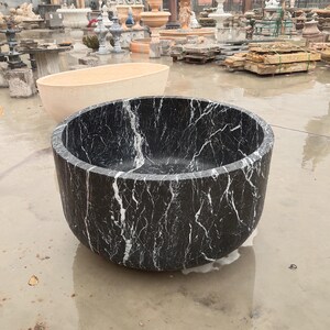 May include: A deep, circular freestanding bathtub carved from black marble with prominent white veining throughout the exterior and interior surfaces. The tub features a smooth, polished finish and a thick, solid wall construction with a rounded base.