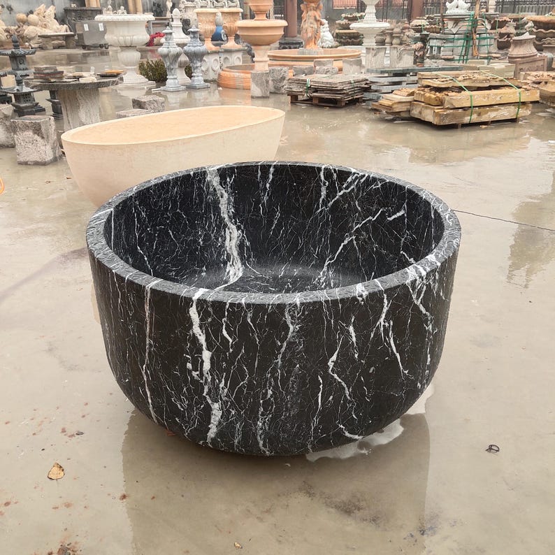 May include: A deep, circular freestanding bathtub carved from black marble with prominent white veining throughout the exterior and interior surfaces. The tub features a smooth, polished finish and a thick, solid wall construction with a rounded base.