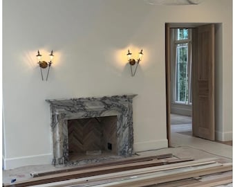 May include: A marble fireplace surround with grey veining features a herringbone brick interior. Two brass wall sconces with dual lights are mounted above the fireplace. A large white pendant light fixture hangs from the ceiling above.