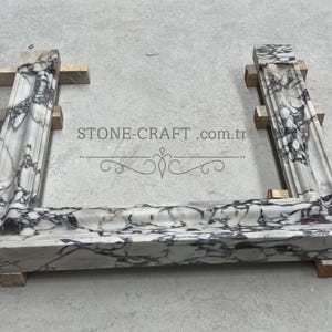 Puede incluir: Un marco de chimenea en forma de U hecho de m&aacute;rmol blanco con vetas pronunciadas de color p&uacute;rpura oscuro y negro. La estructura presenta molduras decorativas y se apoya sobre peque&ntilde;os bloques de madera. El texto STONE-CRAFT.com.tr es visible.