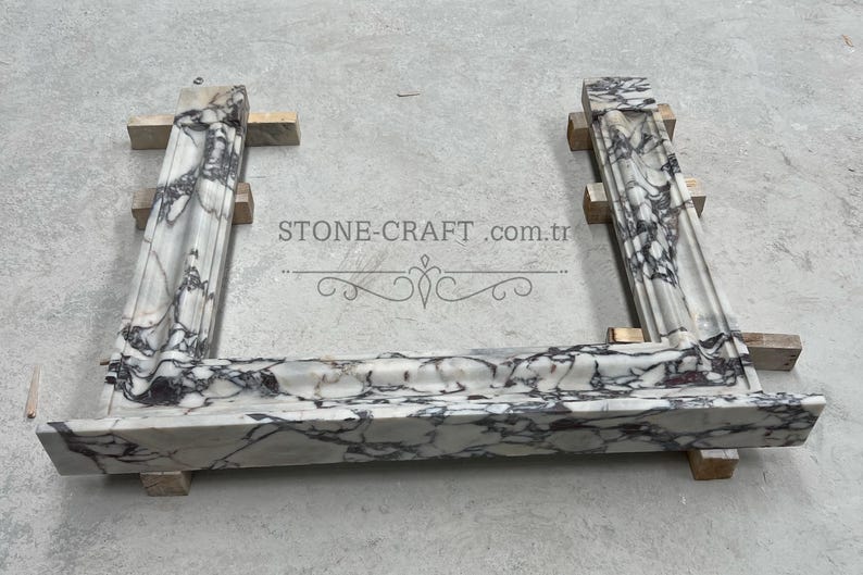 Puede incluir: Un marco de chimenea en forma de U hecho de m&aacute;rmol blanco con vetas pronunciadas de color p&uacute;rpura oscuro y negro. La estructura presenta molduras decorativas y se apoya sobre peque&ntilde;os bloques de madera. El texto STONE-CRAFT.com.tr es visible.